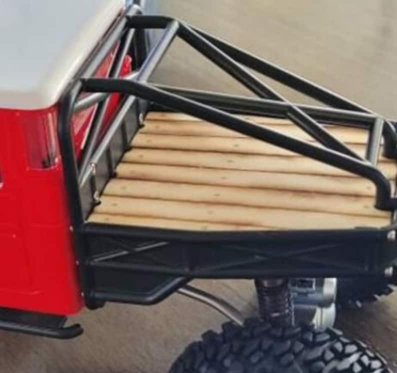 Peut inclure: Gros plan sur un camion jouet rouge et blanc avec une cage de protection noire et une benne en bois. Le camion est &eacute;quip&eacute; de gros pneus noirs tout-terrain et d'une suspension argent&eacute;e.