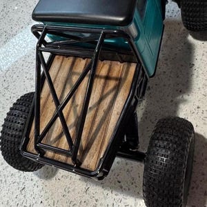 May include: A remote-controlled truck featuring a teal cab with a black roof, a black metal roll cage, and a wooden truck bed. The vehicle is equipped with large, knobby black rubber tires designed for off-road terrain and performance.