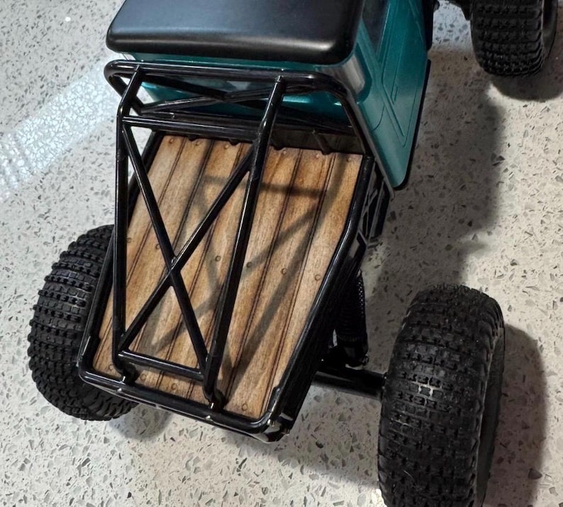 May include: A remote-controlled truck featuring a teal cab with a black roof, a black metal roll cage, and a wooden truck bed. The vehicle is equipped with large, knobby black rubber tires designed for off-road terrain and performance.