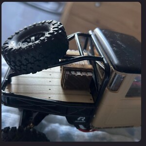 May include: A beige and black remote-controlled truck model featuring a flatbed with a wooden crate and a spare black rubber tire mounted on a black metal roll cage. The truck bed has a light wood-colored surface with visible screw details.