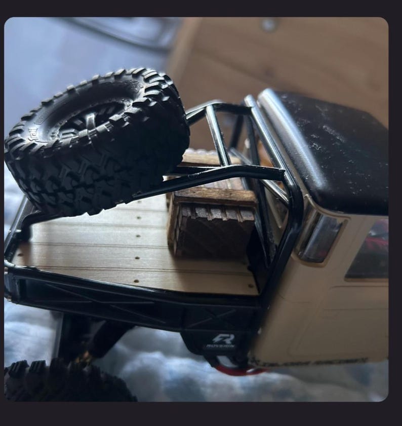 May include: A beige and black remote-controlled truck model featuring a flatbed with a wooden crate and a spare black rubber tire mounted on a black metal roll cage. The truck bed has a light wood-colored surface with visible screw details.
