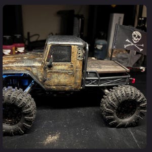 May include: A weathered, gold and black scale model pickup truck with large, knobby off-road tires. The truck features a flatbed with a black flag displaying a white skull and crossbones emblem. The body has a distressed, metallic finish.