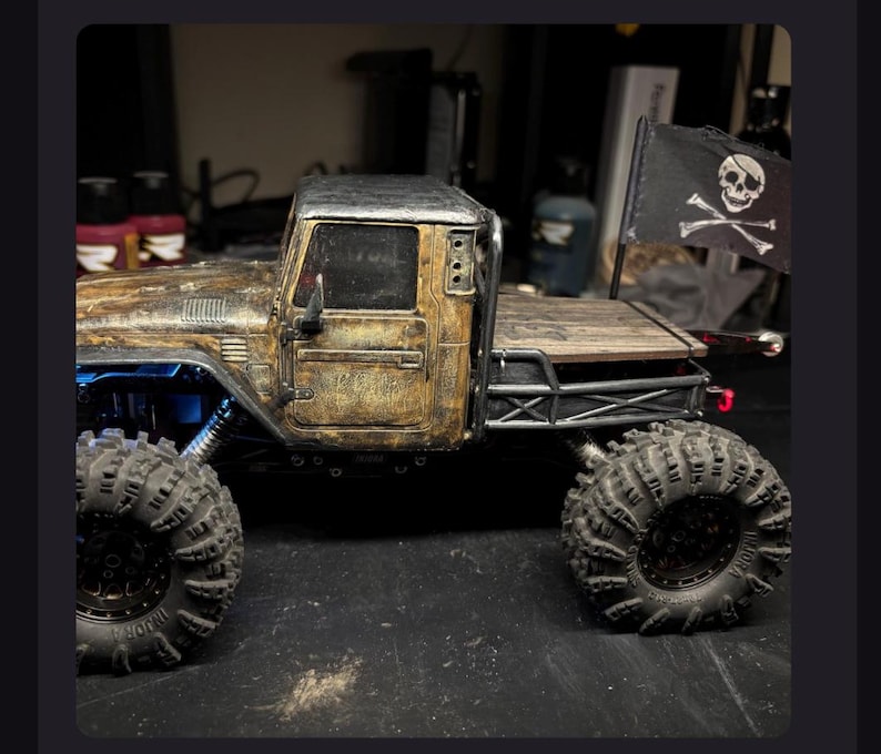 May include: A weathered, gold and black scale model pickup truck with large, knobby off-road tires. The truck features a flatbed with a black flag displaying a white skull and crossbones emblem. The body has a distressed, metallic finish.