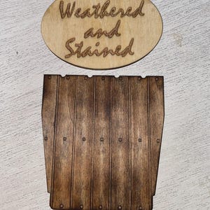 May include: An oval light-colored wooden plaque with the laser-engraved text Weathered and Stained, positioned above a dark brown rectangular wooden piece featuring vertical slats and small circular indentations resembling nail heads.