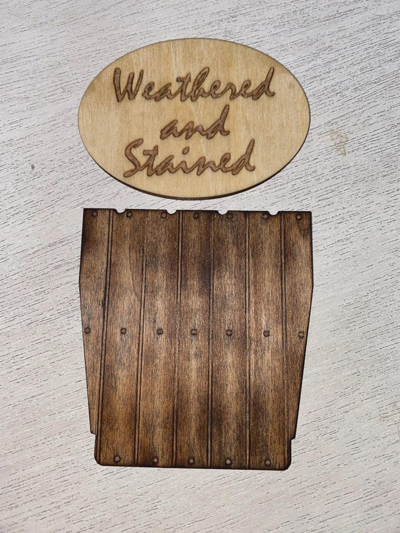 May include: An oval light-colored wooden plaque with the laser-engraved text Weathered and Stained, positioned above a dark brown rectangular wooden piece featuring vertical slats and small circular indentations resembling nail heads.