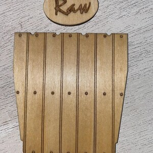 Peut inclure: Une pi&egrave;ce d&eacute;corative en bois brun clair avec le mot "Raw" grav&eacute; sur un ovale. En dessous, une pi&egrave;ce en forme de tonneau, &eacute;galement en bois brun clair, avec des lignes verticales et de petits d&eacute;tails circulaires.