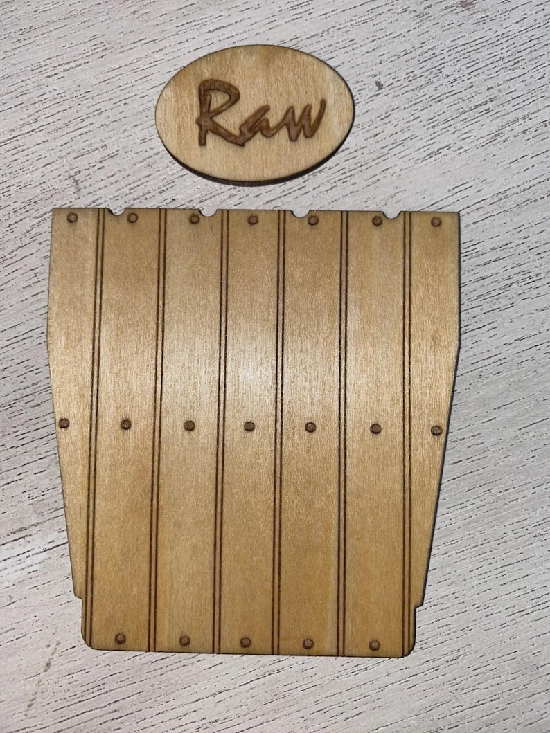Peut inclure: Une pi&egrave;ce d&eacute;corative en bois brun clair avec le mot "Raw" grav&eacute; sur un ovale. En dessous, une pi&egrave;ce en forme de tonneau, &eacute;galement en bois brun clair, avec des lignes verticales et de petits d&eacute;tails circulaires.