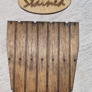 Peut inclure: Panneau en bois avec le mot "Stained" en &eacute;criture cursive au-dessus d'une pi&egrave;ce en forme de tonneau en bois. La pi&egrave;ce en forme de tonneau comporte des lignes verticales et de petits points fonc&eacute;s. Le bois a une teinte brune chaude.