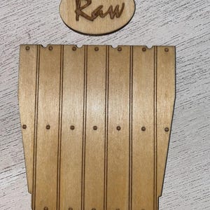 May include: A light brown wooden cutout shaped like a vertical plank wall with engraved lines and small circular dots, accompanied by a separate small oval wooden piece featuring the engraved word Raw in a cursive script font.