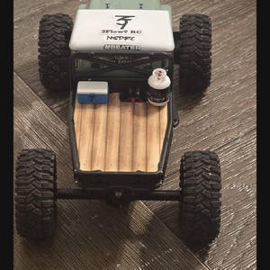 May include: A green remote-controlled truck with a white roof featuring the text 3FLOW9 RC, NSDRC, and #BEATER. The truck bed has a wood-grain floor, a small blue and white cooler, and a miniature white propane tank with black text.