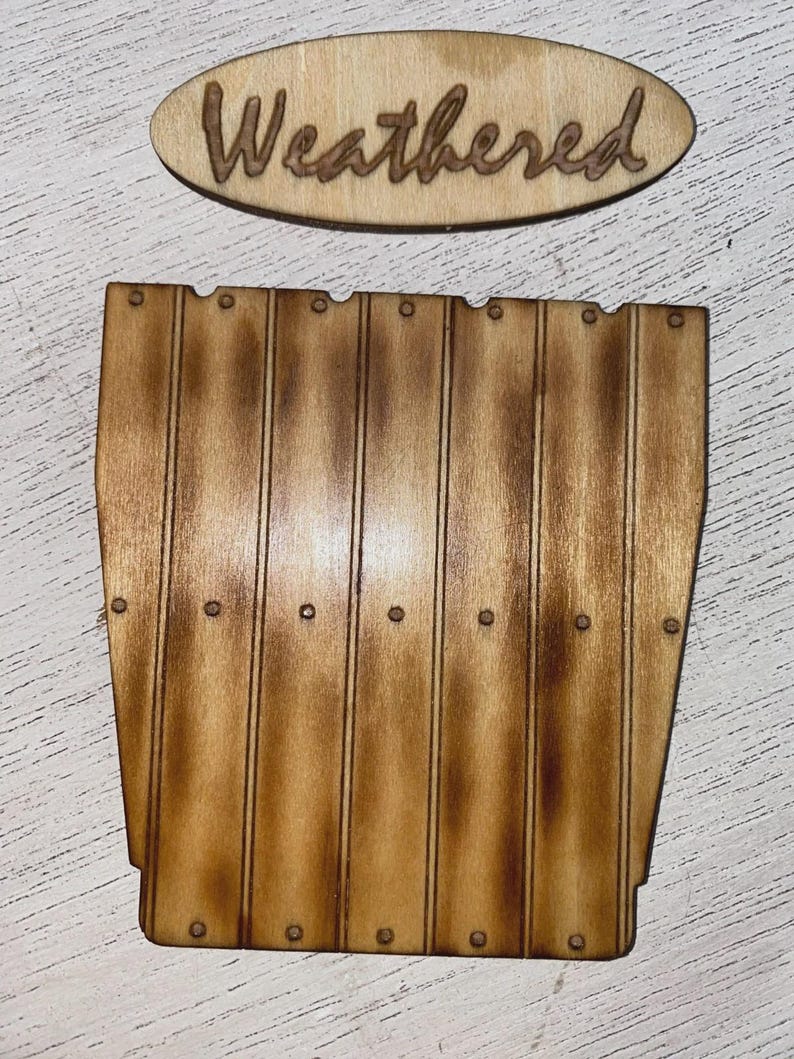 May include: A light brown wooden cutout shaped like a vertical plank wall with dark brown shading and small circular indentations, accompanied by an oval wooden tag featuring the laser-engraved word Weathered in cursive script.