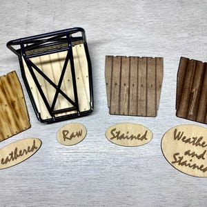 May include: A black metal rack with a light wood base, accompanied by three wooden panels in raw, stained, and weathered finishes. Oval wooden labels are placed below each panel, reading Weathered, Raw, Stained, and Weathered and Stained.