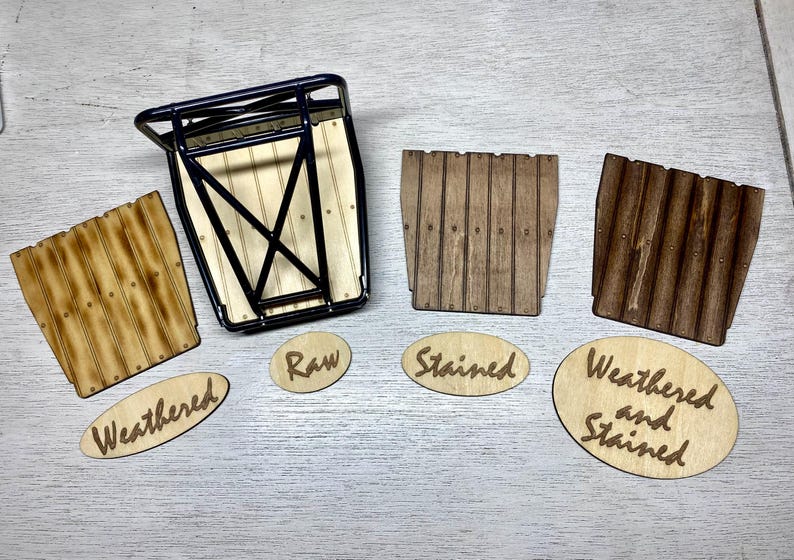 May include: A black metal rack with a light wood base, accompanied by three wooden panels in raw, stained, and weathered finishes. Oval wooden labels are placed below each panel, reading Weathered, Raw, Stained, and Weathered and Stained.