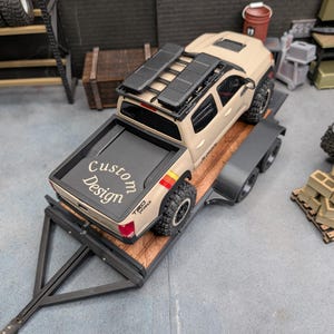 May include: A tan scale model pickup truck with a black roof rack and off-road tires, secured on a flatbed trailer. The truck bed features a black cover with the white text Custom Design printed on it, and red and yellow side decals.