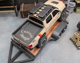 May include: A tan scale model pickup truck with a black roof rack and off-road tires, secured on a flatbed trailer. The truck bed features a black cover with the white text Custom Design printed on it, and red and yellow side decals.