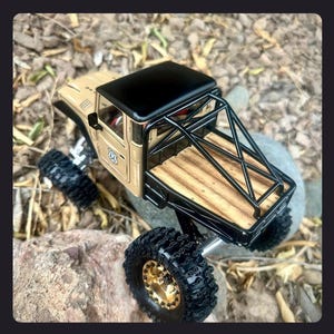 May include: A tan die-cast pickup truck model with a black roof, a black roll cage, and a wooden truck bed. The vehicle features large black off-road tires with gold-colored rims and a small Route 66 emblem on the side door panel.