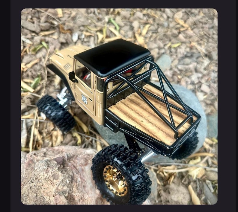 May include: A tan die-cast pickup truck model with a black roof, a black roll cage, and a wooden truck bed. The vehicle features large black off-road tires with gold-colored rims and a small Route 66 emblem on the side door panel.