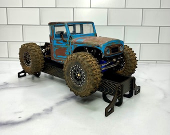 May include: A miniature blue and brown toy truck with large, mud-covered tires. The truck has a weathered appearance, with rust-colored patches on the hood and roof. It sits on a black wooden display stand.