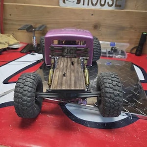 May include: A custom-built, purple-bodied model truck with large, black off-road tires. The truck features a wooden bed, gold-colored shock absorbers, and a detailed chassis. The vehicle is designed for off-road use and is displayed on a red surface.