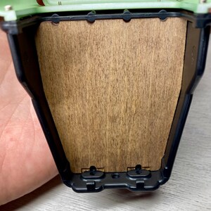 Peut inclure: Gros plan sur un petit conteneur rectangulaire ouvert. Le conteneur a un dessus vert clair et un cadre noir. L'int&eacute;rieur pr&eacute;sente un panneau en bois. Le conteneur est tenu par une main.