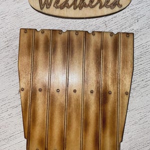 Peut inclure: Panneau en bois avec le mot "Weathered" en &eacute;criture cursive. En dessous, une d&eacute;coupe en bois repr&eacute;sentant une section de cl&ocirc;ture, avec des planches verticales et de petits d&eacute;tails circulaires. Le bois a une teinte marron chaude.