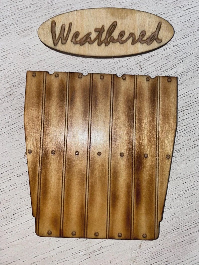 Peut inclure: Panneau en bois avec le mot "Weathered" en &eacute;criture cursive. En dessous, une d&eacute;coupe en bois repr&eacute;sentant une section de cl&ocirc;ture, avec des planches verticales et de petits d&eacute;tails circulaires. Le bois a une teinte marron chaude.