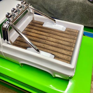May include: A white toy pickup truck with a wooden bed and chrome roll bar. The truck sits on a bright green surface. The bed of the truck has a dark wood grain pattern.