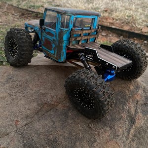 May include: A remote-controlled truck model with a weathered blue cab, rust-colored accents, and a wooden flatbed. It features large, knobby black tires with beadlock wheels, blue metal suspension components, and a small MOFO RC sticker on the rear window.
