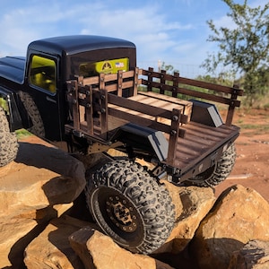 May include: A black remote-controlled pickup truck featuring a dark wood stake bed with vertical slats and a matching wooden floor. The truck has yellow-tinted windows, large black off-road tires, and a small sticker on the rear window.