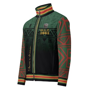 Op de afbeelding: Een groene, zwarte en rode trainingsjas met ritssluiting en een hoge kraag. De jas heeft geometrische patronen, de tekst "M.L.K. 1963" en de zin "I have a dream". De jas heeft een gouden rits en afwerking.