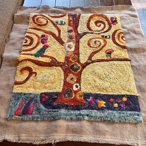 May include: A rectangular rug hooking piece featuring a central tree with swirling branches in shades of yellow, orange, and brown. The design includes small birds, floral motifs, and a dark blue base with colorful accents, all worked on a natural burlap backing.