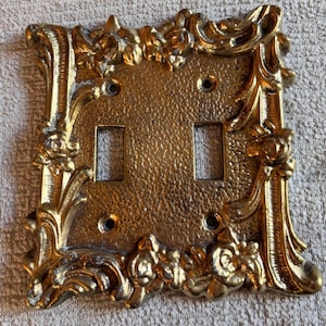 May include: A gold-colored metal double light switch wall plate featuring a textured center panel and an ornate, raised border with floral and scrollwork details. The rectangular plate includes two rectangular cutouts and four screw holes.