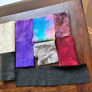 May include: A collection of eight rectangular fabric swatches in various colors and textures, including cream, purple, tie-dye blue and pink, dark maroon, light gray, bright red, and two pieces of dark charcoal gray felted material.