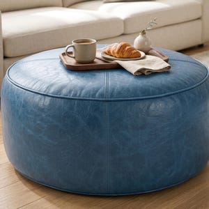 May include: A round, blue faux leather ottoman with visible stitched seams. On top sits a rectangular wooden tray holding a beige ceramic mug filled with coffee, a small white vase with a sprig, a croissant on a plate, and a folded linen napkin.