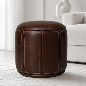 May include: A cylindrical ottoman upholstered in dark brown leather featuring a smooth, rounded top surface and vertical paneling along the sides. The structure is accented with visible white contrast stitching along the seams of each panel.