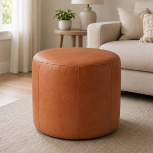 May include: A round, tan-colored ottoman with a smooth, solid surface and vertical seam detailing. The piece features a cylindrical shape and a consistent, matte-textured finish, providing a stable and uniform appearance for use as a footrest.
