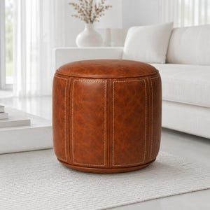 May include: A round, cognac-colored faux leather ottoman featuring vertical paneling with visible white stitching. The cylindrical footstool has a smooth, flat top surface and a structured base, showcasing a consistent textured finish throughout.