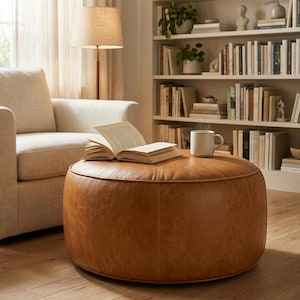 Peut inclure: Un pouf rond en cuir beige avec des coutures visibles sur le bord supérieur. Un livre ouvert aux pages blanches et une tasse en céramique de couleur claire sont posés sur la surface plane du pouf, qui repose sur un plancher en bois clair.