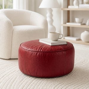 May include: A round, deep red leather ottoman with visible stitched seams. A white hardcover book and a light-colored ceramic mug are placed on top of the ottoman. The surface of the leather has a textured, slightly wrinkled appearance.