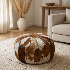 May include: A round, low-profile ottoman constructed from stitched panels of genuine cowhide. The surface features a patchwork of natural fur in varying shades of brown, tan, and white, arranged in a radial pattern meeting at a central circular piece.
