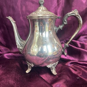 May include: A silver-colored teapot with ornate detailing, including a decorative handle and spout. The teapot has a lid with a floral finial and rests on three decorative feet. The polished surface reflects the surrounding environment.