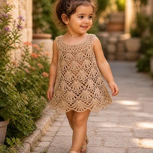 May include: A little girl wearing a sleeveless, tan crochet dress with a scalloped hemline. The dress has a detailed, openwork pattern. She is also wearing tan sandals. The background shows a stone walkway and greenery.