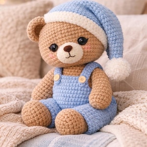 May include: A handmade crochet teddy bear wearing a light blue nightcap with a white brim and pom-pom. The bear has a light brown body, rosy cheeks, and is dressed in blue overalls with wooden buttons. The bear is sitting on a bed with soft, neutral-colored blankets.