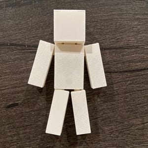 Könnte beinhalten: Eine weiße, blockartige Figur mit rechteckigem Kopf, Rumpf, Armen und Beinen. Die Arme und Beine sind am Rumpf befestigt. Die Figur besteht aus einem festen, undurchsichtigen Material und befindet sich auf einer dunkelbraunen Holzoberfläche.