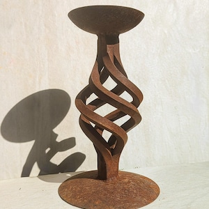 May include: A rust-colored metal candle holder featuring a circular base, a central stem with a twisted, open-work spiral design, and a wide, bowl-shaped top platform. The surface of the object has a textured, weathered appearance throughout.