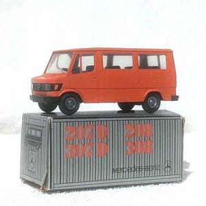 May include: An orange Mercedes-Benz van toy model resting on its original rectangular cardboard box. The box features vertical gray stripes, the Mercedes-Benz logo, and the printed text 207D, 208, 307D, and 308 in orange lettering.