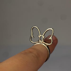 May include: A silver-toned metal ring featuring a decorative bow-shaped element on the top. The bow design is open and wire-like, with a small rectangular hallmark visible at the base where the bow connects to the circular band of the ring.