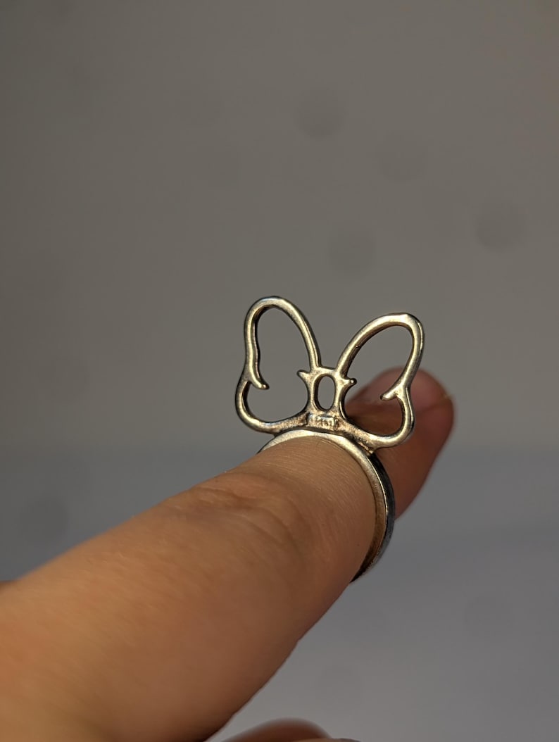 May include: A silver-toned metal ring featuring a decorative bow-shaped element on the top. The bow design is open and wire-like, with a small rectangular hallmark visible at the base where the bow connects to the circular band of the ring.
