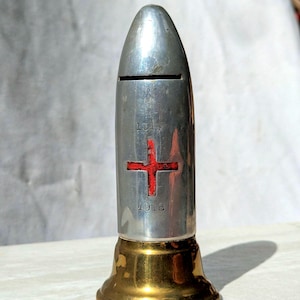Puede incluir: Una hucha de metal plateado con forma de bala y una ranura horizontal cerca de la parte superior. El cuerpo presenta un emblema de cruz roja centrado entre los años grabados 1914 y 1918. El objeto se apoya sobre una base de metal color latón.