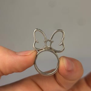 May include: A silver-toned metal ring featuring a cutout bow design on the top. The center of the bow includes a small, oval-shaped opening. The word Disney is stamped in small, clear letters at the base of the bow where it meets the ring band.